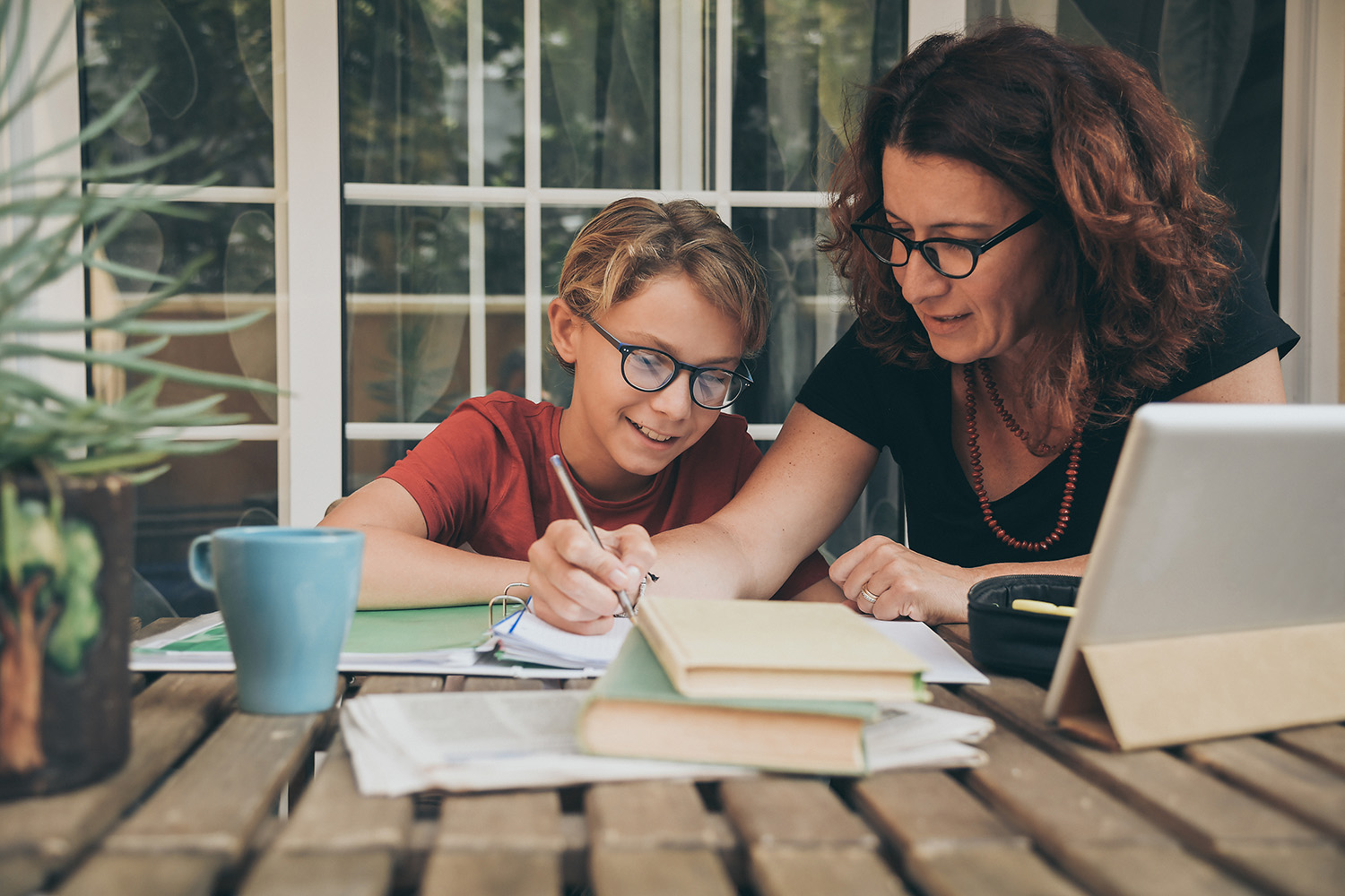 Young student doing homework at home with school books, newspaper and digital pad helped by his mother. Mum writing on the copybook teaching his son. Education, family lifestyle, homeschooling concept